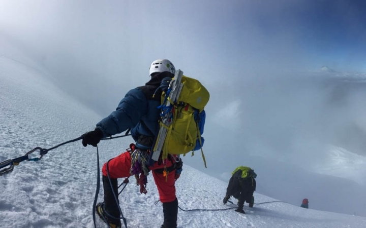 A group of mountaineers wearing safety gear are connected by a rope. The person in the foreground looks down the snowy slope to two others below them.